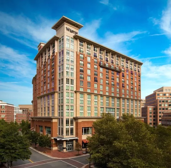 A tall, modern hotel building with Westin signage on top, featuring large windows and red-brick accents, stands on a city corner surrounded by trees under a bright blue, lightly clouded sky.