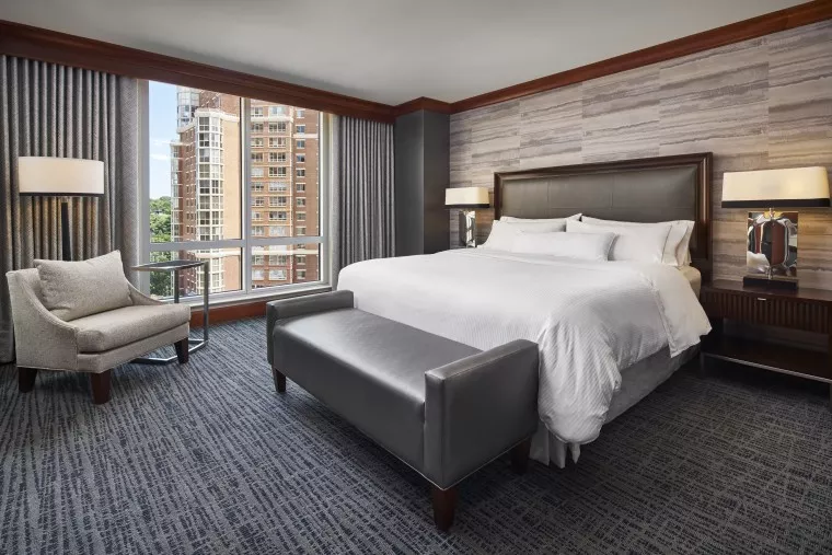 Modern hotel room with a large bed, upholstered bench, armchair, side tables with lamps, and a window showing tall buildings outside. The decor features neutral tones and patterned wallpaper.