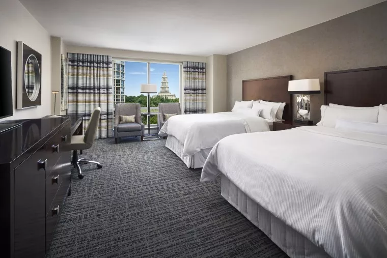 A modern hotel room with two large beds, a work desk and chair, armchairs by a window, dark wood furniture, and a view of a building with a spire outside.