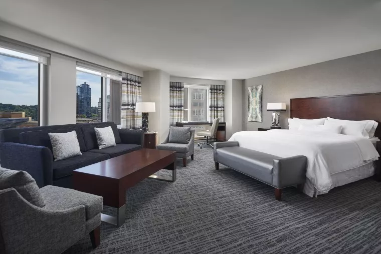 Spacious hotel room with a king-size bed, gray upholstered bench, dark wood headboard, seating area with sofa and armchairs, coffee table, desk, lamps, and large windows with city views.