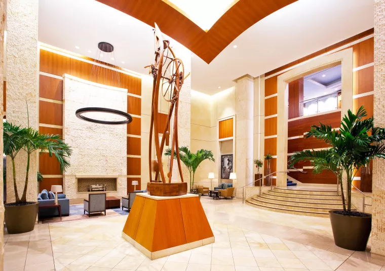 A spacious, modern hotel lobby with tall ceilings, large potted plants, sleek wood and stone finishes, contemporary art sculpture in the center, and seating areas with sofas around a fireplace.