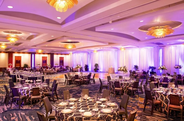 A spacious, elegantly decorated banquet hall with round tables set for an event, featuring patterned tablecloths, floral centerpieces, dim lighting, and a large dance floor with purple uplighting.