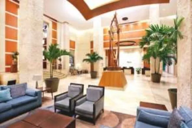 Spacious hotel lobby with modern decor, tall marble columns, large potted plants, comfortable seating, and a central abstract sculpture. Warm lighting and high ceilings create an inviting atmosphere.