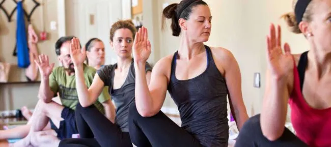 A group of people practice yoga indoors, sitting on mats in a twist pose with one hand raised and looking over their shoulders. They appear focused and are wearing athletic clothing.