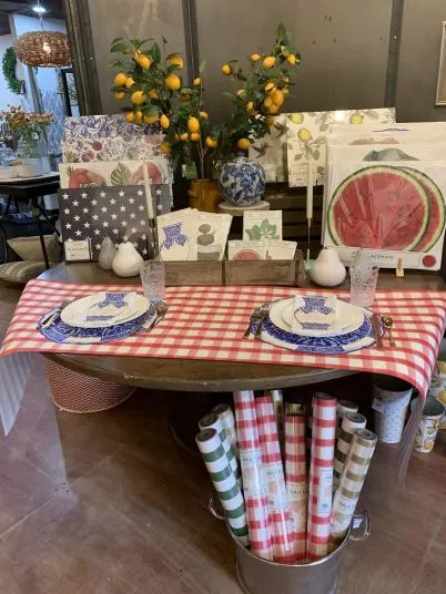 A table set for two with blue and white dishes on a red checkered tablecloth, surrounded by rolls of wrapping paper, watermelon napkins, and lemon decorations. Lemon branches and various tableware are displayed in the background.