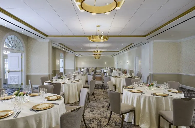 Elegant banquet hall with round tables covered in white cloths, gold plates, glasses, and simple floral centerpieces. Neutral-toned chairs surround the tables, and modern chandeliers hang from the ceiling. Large windows line the walls.