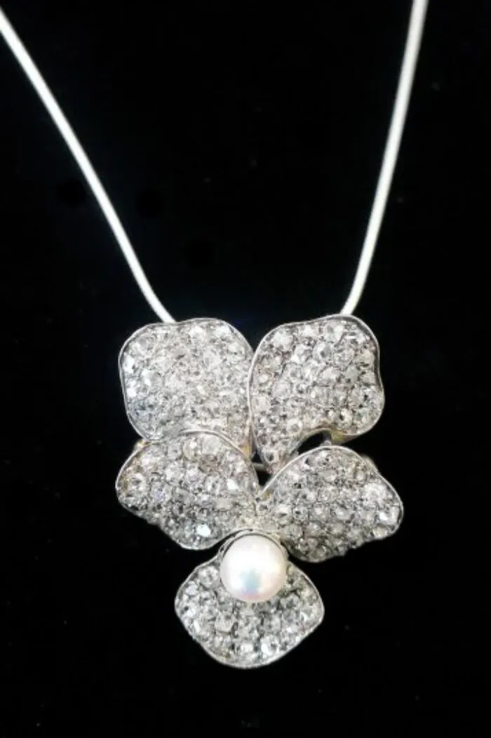 A silver necklace with a pendant shaped like a flower, featuring sparkling crystals on the petals and a round white pearl at the center, displayed against a black background.