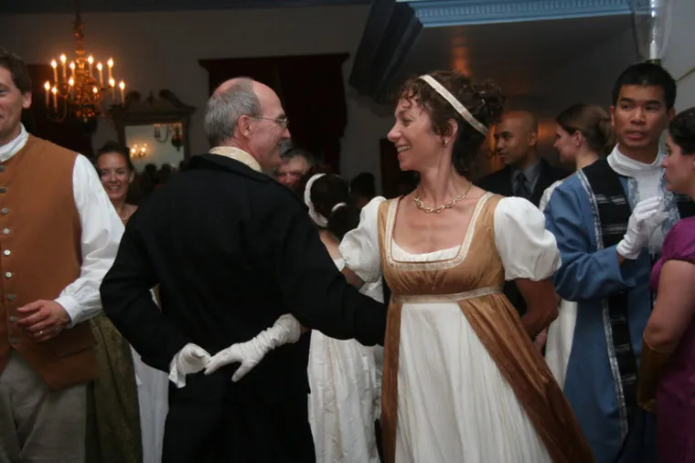 A group of people in historical costumes dance at a formal indoor event. A woman in a brown Regency-style dress smiles at a man in a black coat and white gloves as they dance together. Other guests mingle in the background.