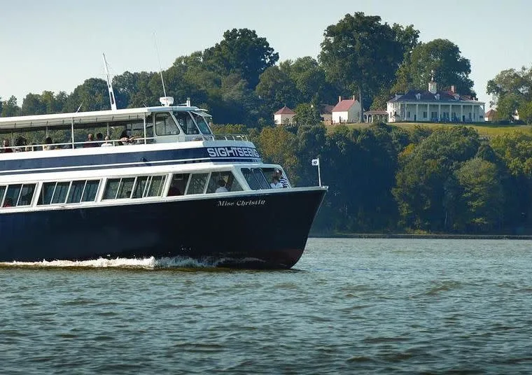 A sightseeing tour boat cruises along a river, with a large historic white mansion surrounded by trees visible on a hill in the background.