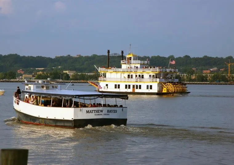 Two boats travel on a river: a white paddle steamboat with a yellow paddlewheel in the background, and a smaller boat named Matthew Hayes with passengers in the foreground. Trees and buildings line the shore.