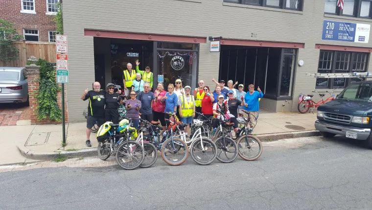 A group of cyclists wearing helmets and colorful biking gear pose with their bikes on a sidewalk in front of a brick building. Some are smiling and waving at the camera. A few bikes are parked along the curb.