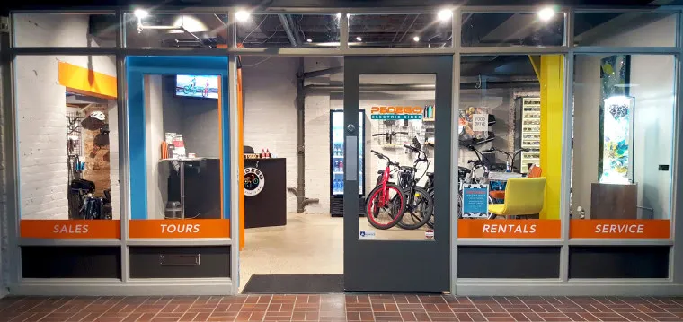 Storefront of a bike shop with large glass windows and door, displaying bicycles inside. Orange signs read Sales, Tours, Rentals, and Service. The shop is well-lit, modern, and organized.