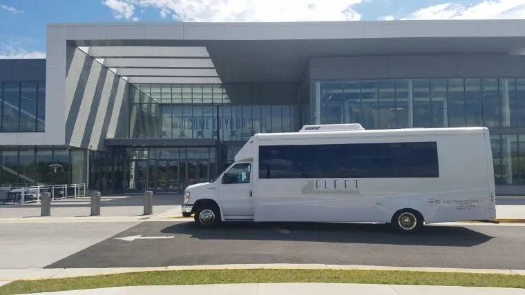 A white shuttle bus labeled Fleet Transportation is parked outside a modern glass building entrance on a sunny day. The building features large windows and a welcoming sign.