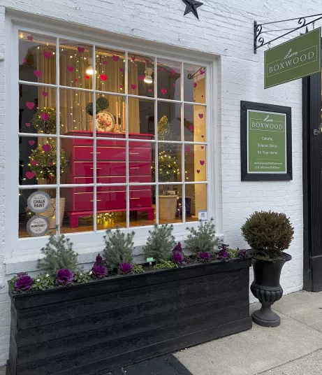 A shop window display with a bright pink dresser, string lights, heart decorations, small trees, and potted plants. The sign above reads Boxwood. The exterior features white brick and a black planter with greenery.