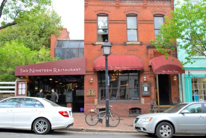 A red-brick building with maroon awnings houses Two Nineteen Restaurant. Two cars are parked out front, and a bicycle is locked to a lamp post on the sidewalk. Trees line the left side of the building.