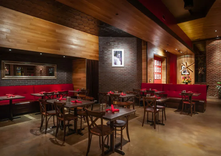 A stylish restaurant with wooden tables and chairs, red cushioned seating along brick walls, dim lighting, and contemporary decor. Red napkins are neatly placed on each table, creating a warm, inviting atmosphere.