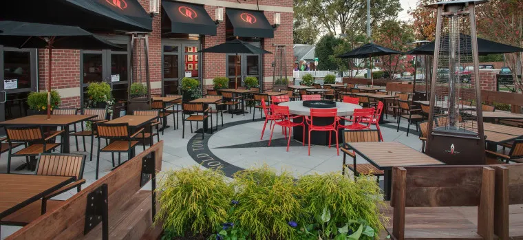 Outdoor restaurant patio with wooden tables and chairs, some grouped around a circular fire pit. Red chairs surround the fire pit, and black umbrellas provide shade. Green plants and flowers frame the seating area.