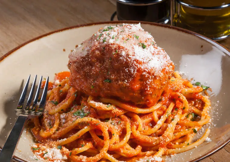 A plate of spaghetti topped with tomato sauce, a large meatball, and grated cheese, with a fork resting on the side. The background shows a wooden table and glass containers.