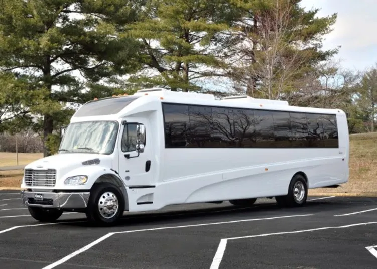 A large white shuttle bus is parked in an empty lot with trees and grass in the background on a clear day.