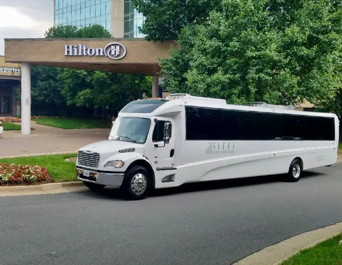 A white shuttle bus is parked on a road in front of the Hilton hotel entrance, with trees and landscaping visible in the background.