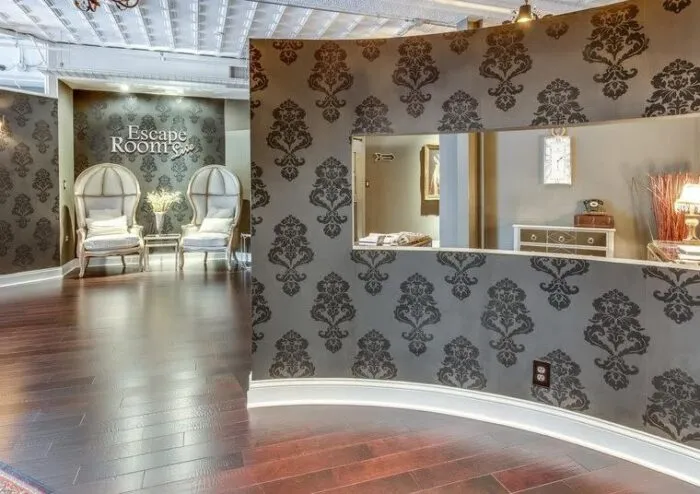 Elegant escape room lobby with dark floral wallpaper, two large high-backed chairs, a small table, and a decorative reception area with a large cutout window. The floor is dark wood, creating a sophisticated atmosphere.