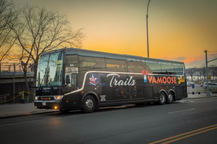 A black Vamoose Trails coach bus is parked on a city street at sunset, with bare trees and a fence in the background. The sky is orange and yellow, creating a warm glow over the scene.