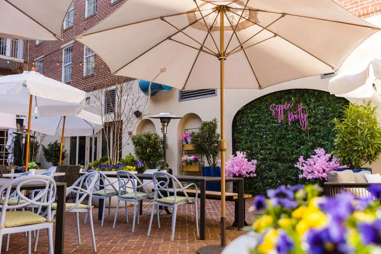 An outdoor patio with tables and chairs under large white umbrellas. Pink and purple flowers decorate the area, and a neon sign reading Sip n Party hangs on a green wall in the background.