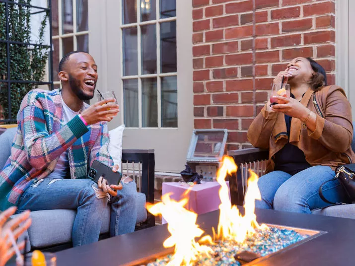 Two people sit on outdoor couches by a fire pit, laughing and enjoying drinks. They appear relaxed and happy, surrounded by a cozy atmosphere with brick walls and large windows in the background.