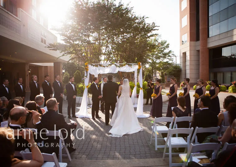 An outdoor wedding ceremony with a bride and groom standing under a decorated canopy, surrounded by bridesmaids in dark dresses and groomsmen in tuxedos, while guests watch from white chairs.