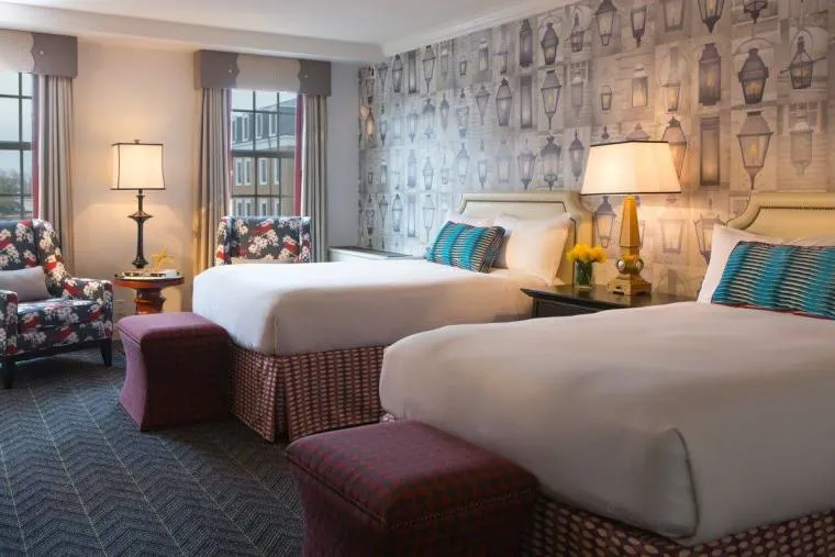 Hotel room with two double beds, patterned headboards, and blue accent pillows. The wall features lantern-themed wallpaper. There are two red ottomans, floral armchairs, a lamp, and large windows with gray curtains.