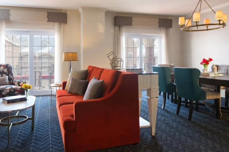 A bright living and dining area with a red sofa, blue chairs around a dining table, large windows letting in natural light, and modern decor elements.