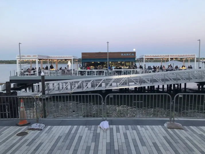People dine outdoors at a waterfront restaurant called BARCA on a pier, with railings and a walkway in the foreground and calm water in the background at dusk.