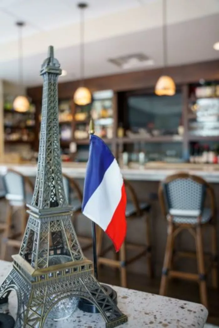 A small Eiffel Tower model and a French flag are displayed on a counter in a restaurant or café, with a bar and high chairs visible in the blurred background.