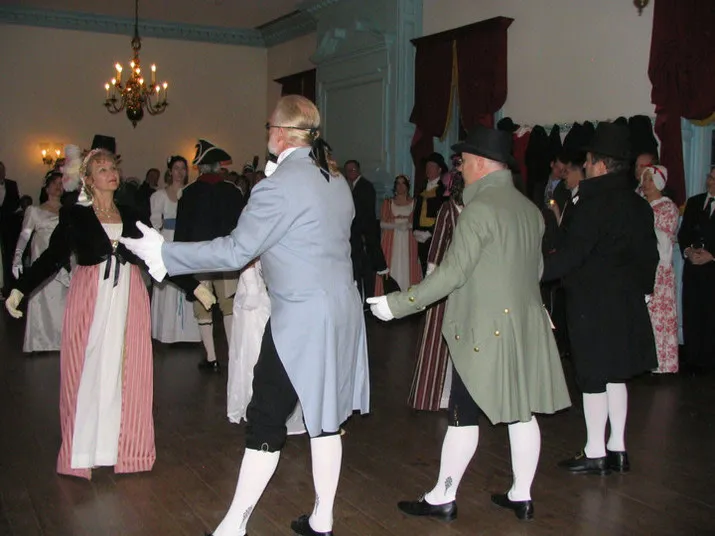 People wearing historic 18th- or 19th-century costumes dance in a ballroom with chandeliers and wooden floor. The scene includes women in long dresses and men in coats, breeches, and hats.