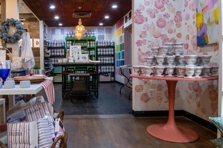 A colorful boutique filled with home goods, featuring stacked bowls on a round table, striped linens, glassware, and shelves lined with bottles and jars against patterned and vibrant walls.
