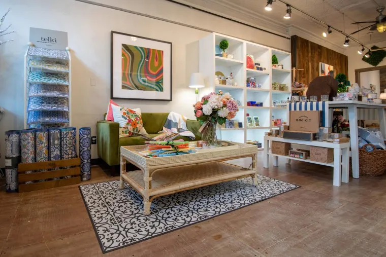 A cozy boutique shop with a green armchair, a coffee table with flowers, colorful decor, shelves filled with gifts, and bright lighting. The atmosphere is inviting and stylish, with patterned rugs and wall art.