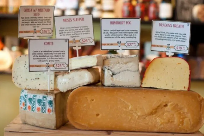 A display of various cheeses on a wooden board, each with a label describing its name, price, and details. The cheeses are different shapes and colors, with rind and creamy textures visible. A blurred shop background is behind them.
