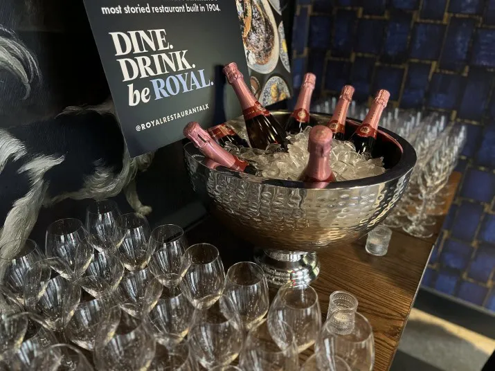 A large silver bowl filled with ice and pink-capped bottles of sparkling wine sits beside rows of empty wine glasses and a sign that reads, DINE. DRINK. be ROYAL.