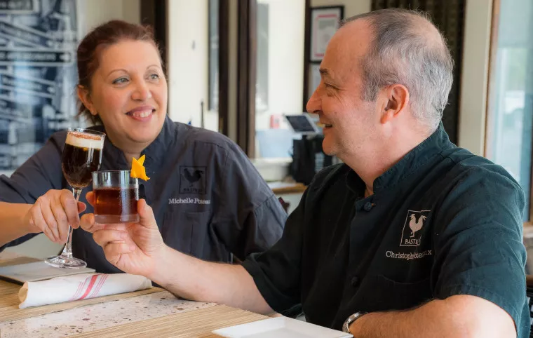 Two smiling chefs sit at a table, toasting drinks. The woman holds a glass with frothy topping, the man holds a cocktail with an orange garnish. Both wear dark chef jackets with embroidered names.