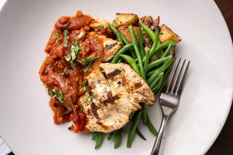 A plate with grilled chicken breast, green beans, roasted potatoes, and tomato sauce topped with fresh herbs, with a fork resting on the plate.