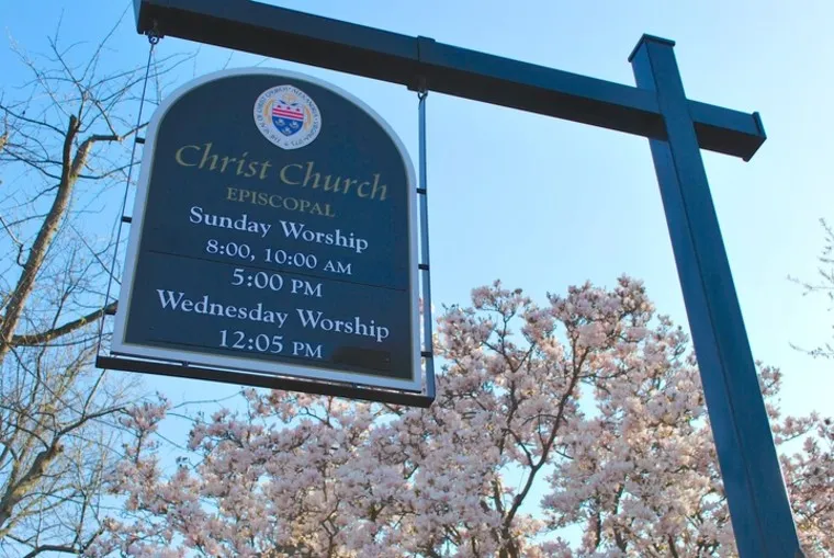 A sign for Christ Church Episcopal lists Sunday worship at 8:00, 10:00 AM, and 5:00 PM, and Wednesday worship at 12:05 PM. The sign hangs outdoors near blooming trees under a clear sky.