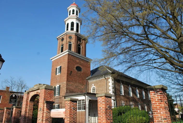 A historic red brick church with a tall white-steepled bell tower stands behind a brick fence and iron gate, surrounded by trees under a clear blue sky.