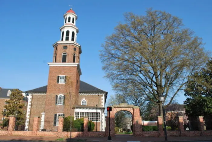 A historic brick church with a tall, white cupola stands behind a brick fence and archway. A large tree with sparse leaves is on the right, and the sky is clear and blue.