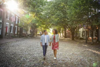 A couple holds hands while walking down a sunlit, tree-lined cobblestone street in a residential neighborhood with American flags on houses.