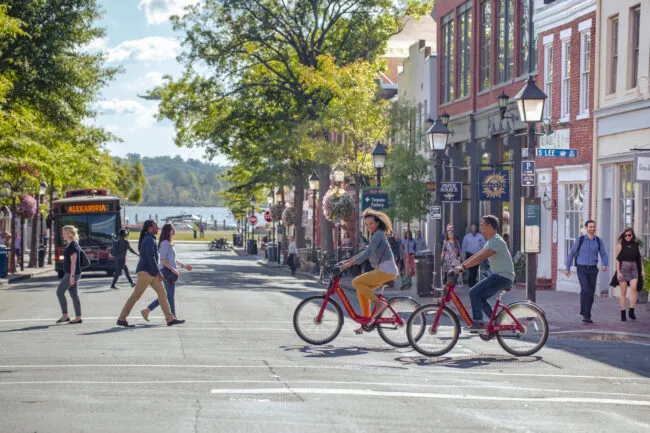 People cross a sunny, tree-lined street in a lively urban area. Two people ride red bikes, others walk, and a city bus approaches in the background. Historic brick buildings line the street.