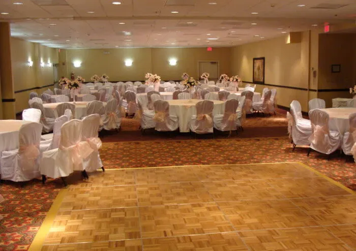 A banquet hall with round tables covered in white cloths and chairs with white covers and pink sashes, arranged around a wooden dance floor. Floral centerpieces are on each table. The room is softly lit.