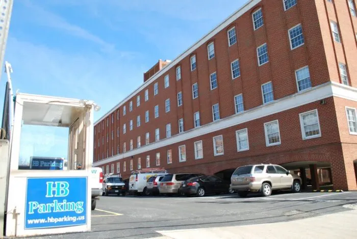 A parking lot next to a multi-story red brick building, with several parked cars and a small booth displaying an HB Parking sign in the foreground.