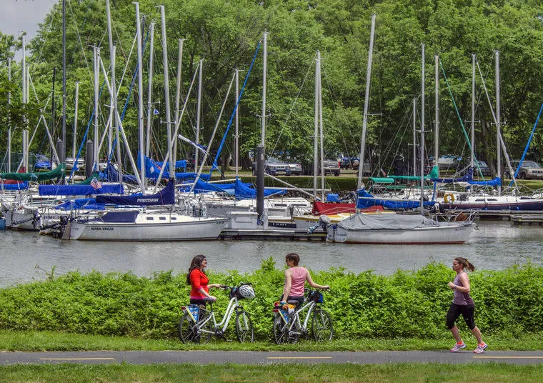 Two people stand with bicycles talking while another person jogs past them on a path, with numerous sailboats docked in a marina and green trees in the background.