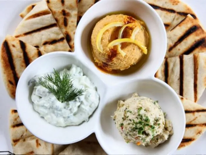 A plate with grilled pita bread slices arranged around a divided dish containing hummus with lemon slices, tzatziki garnished with dill, and a scoop of baba ganoush with herbs.
