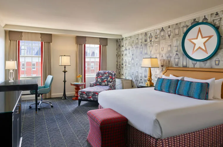 A hotel room with a large bed, patterned wallpaper, a star-shaped wall decor, an armchair, a desk, two windows with drapes, and various lamps, creating a cozy and stylish atmosphere.